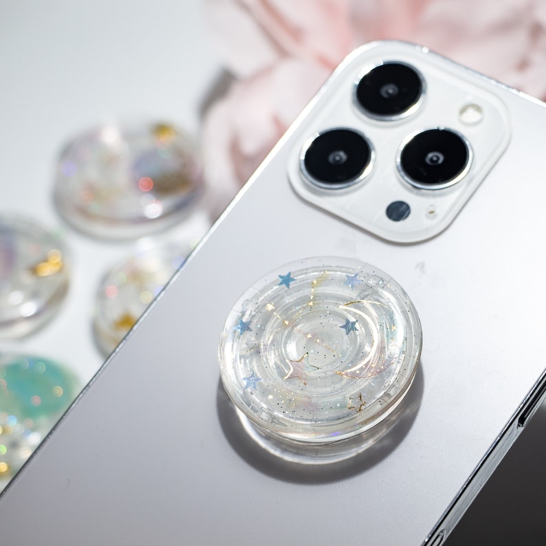 May include: A clear phone grip with a swirling design and gold glitter. The grip has blue stars and a crescent moon shape. The grip is on a white iPhone.