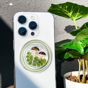 May include: A white iPhone case with a clear, round phone grip featuring a small, dried fern and two brown mushrooms.