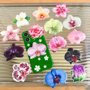May include: A green phone case with white flower accents and a large pink orchid design, surrounded by various colourful orchid-shaped phone grips. The grips feature shades of pink, purple, white, and blue, with intricate details.