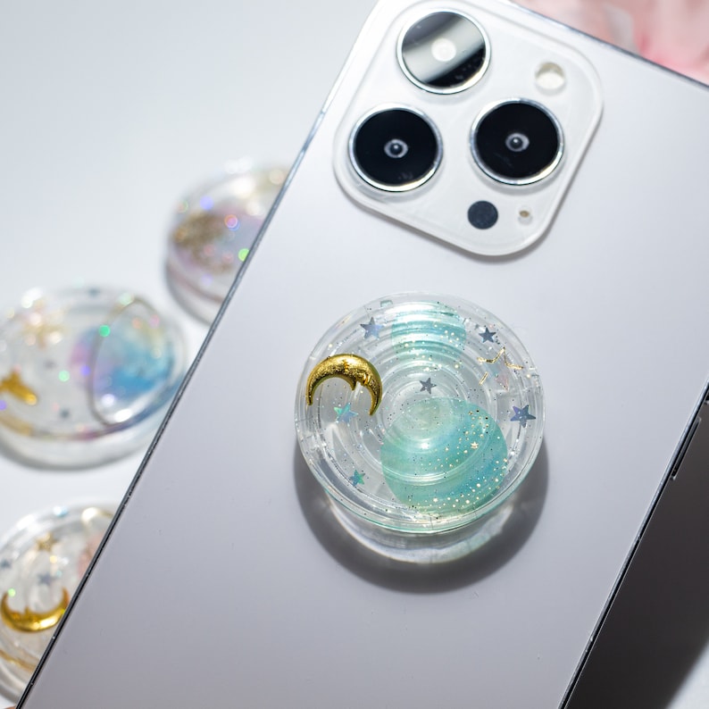 May include: A clear phone grip with a blue and green swirl design. The grip has a gold crescent moon and silver glitter stars embedded in the resin.