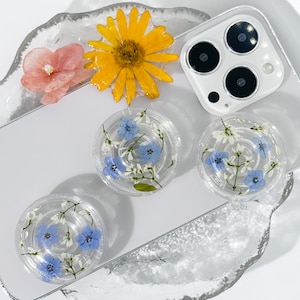 May include: Three clear resin phone case charms featuring pressed blue and white flowers. The charms are round and have a white background with blue flowers and white stems.