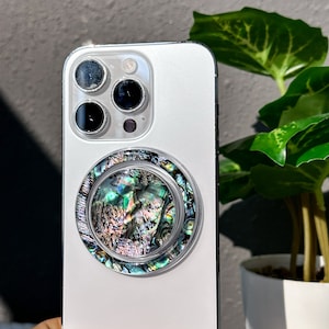 Puede incluir: Un iPhone blanco con un agarre de teléfono de concha negra e iridiscente. El agarre tiene un diseño circular elevado.