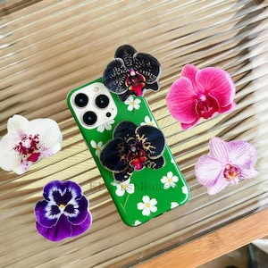 Może przedstawiać: Zielone etui na telefon z białym wzorem stokrotek i dołączonymi czarnymi kwiatami orchidei. Dodatkowe kwiaty orchidei w kolorze różowym, fioletowym i białym są rozrzucone wokół etui.