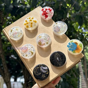 Pode incluir: Uma coleção de nove suportes circulares para telemóvel dispostos numa superfície bege. Os designs incluem peças de resina transparente com flores vermelhas ou amarelas, motivos celestiais com luas e estrelas douradas e peças pretas.