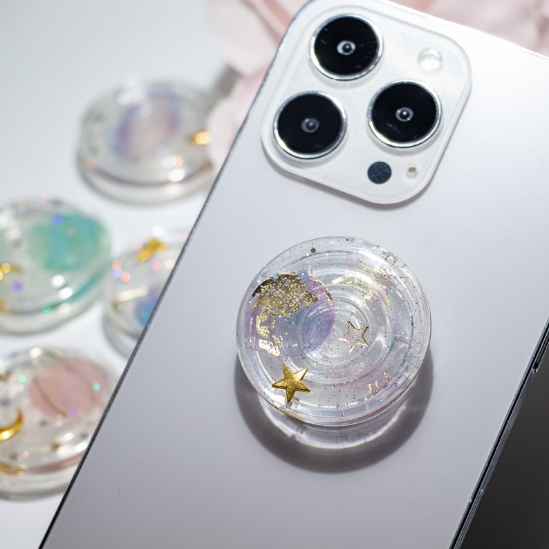 May include: A clear phone grip with a swirling galaxy design and gold glitter. The grip has a raised circle in the center and two gold stars.