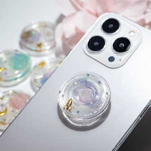 May include: A white phone case with a clear phone grip featuring a galaxy design with a gold rocket and silver glitter.