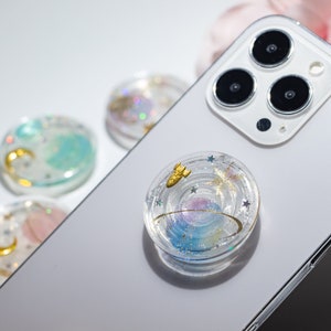 May include: A clear phone grip with a galaxy design. The grip features a gold rocket, stars, and a swirl pattern. The grip is on a white phone.