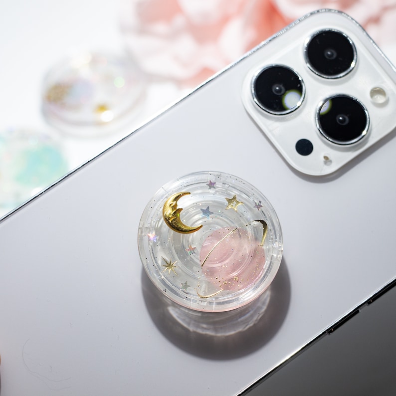 May include: A clear phone grip with a pink and gold galaxy design. The grip has a crescent moon, a planet, and stars.