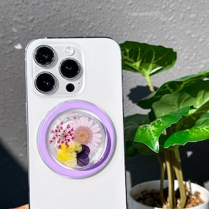 Puede incluir: Una funda blanca para iPhone con un soporte de anillo morado que presenta flores prensadas. Las flores son una mezcla de rosa, amarillo y morado.