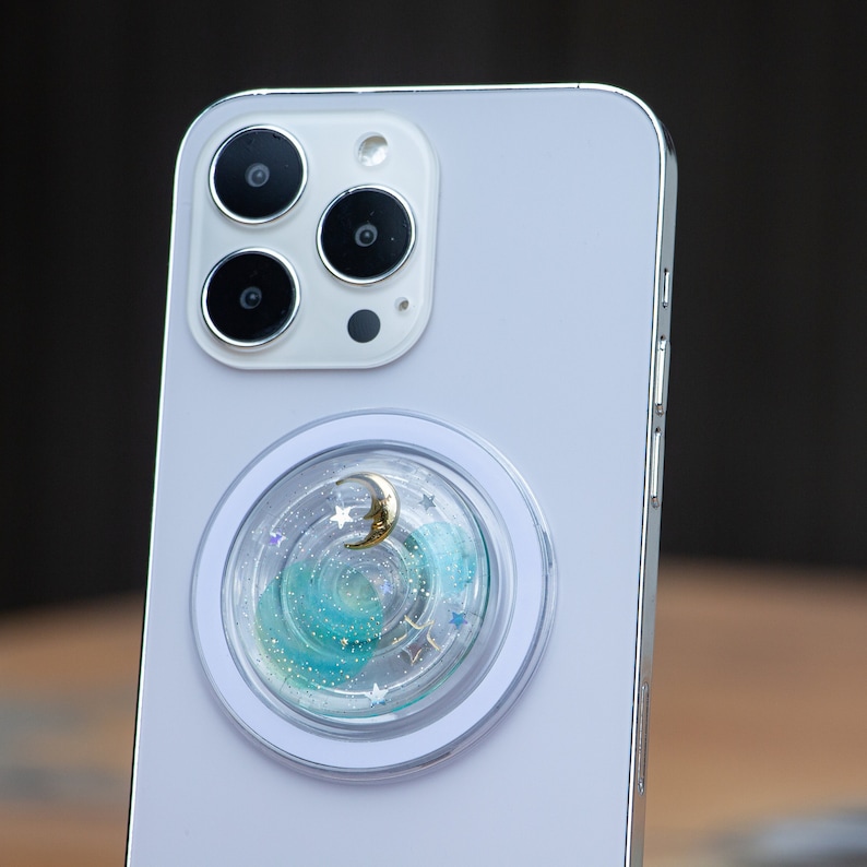 May include: A white smartphone with a silver frame and a clear phone grip with a blue and green glitter design and a gold crescent moon charm.
