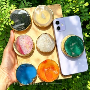 May include: A collection of six circular phone grips featuring polished agate slices in black, beige, pink, white, blue, and orange, each with a gold-toned metallic edge. One green agate phone grip is attached to a light purple smartphone.