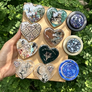 May include: A collection of heart-shaped and circular phone grips featuring diverse designs including cat portraits, floral patterns, iridescent shell fragments, and celestial charms. Some grips display text, while others showcase metallic or swirling blue finishes.
