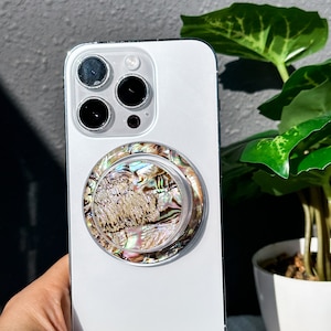 Op de afbeelding: Een witte iPhone met een regenboogkleurige schelptelefoonhouder bevestigd aan de achterkant. De telefoon heeft een drievoudig camerasysteem.