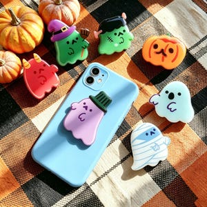 Pode incluir: Uma capa de smartphone azul-clara com um fantasma roxo usando um chapéu verde, rodeada por vários suportes de telefone com tema de Halloween, incluindo um fantasma bruxo verde, um monstro verde, uma abóbora laranja, um diabo vermelho e uma múmia.