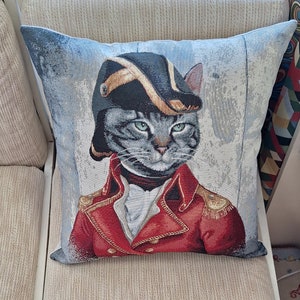 Puede incluir: Una almohada decorativa con un gato gris que lleva un uniforme militar rojo y dorado y un sombrero negro. El gato tiene una expresión seria y mira directamente al espectador.