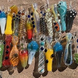 Könnte beinhalten: Eine Sammlung farbenfroher Glaspfeifen mit verschiedenen Designs. Die Pfeifen weisen Wirbel, Punkte und andere dekorative Elemente in Blau-, Gelb-, Rot- und Grüntönen auf. Einige haben kleine, dekorative Glasperlen.
