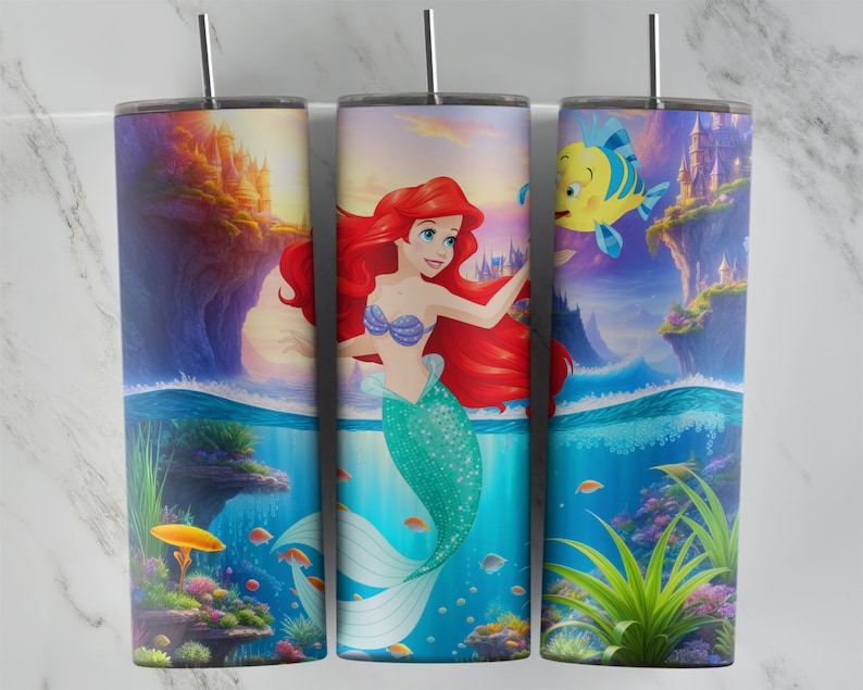 K&ouml;nnte beinhalten: Drei Edelstahlbecher mit Meerjungfrauen-Motiv. Die Becher zeigen eine farbenfrohe Unterwasser-Szene mit Arielle, Flounder und einem Schloss im Hintergrund.