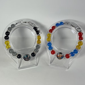 May include: Two beaded bracelets featuring circular charms. The left bracelet has black, yellow, and gray beads with a Batman charm. The right bracelet has red, yellow, and blue beads with a Superman charm. Both include silver-toned metal spacers.