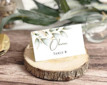 May include: A white place card with the name "Olivia" and "Table 8" in elegant script, adorned with watercolor-style green leaves and gold accents. The card rests on a rustic wooden slice, suggesting a wedding or event theme.