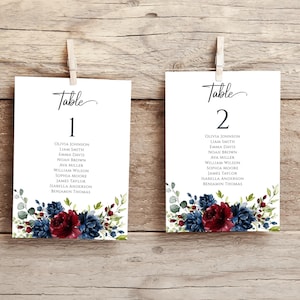 May include: Two white cards with black text and floral designs. The cards are labeled "Table 1" and "Table 2" and list names for each table. The floral designs are in shades of blue, green, and burgundy.