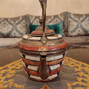 May include: A decorative teapot with a brass handle and spout. The body features a geometric pattern of white, orange, and brown segments. The teapot sits on a patterned tablecloth, suggesting a cultural or vintage theme.