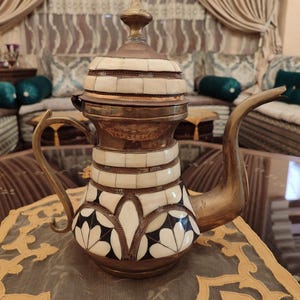 May include: A decorative brass teapot with a white and black geometric pattern. The teapot has a lid with a finial and a spout. The teapot is sitting on a table with a lace tablecloth.