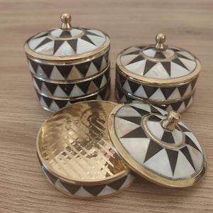 Op de afbeelding: Drie ronde doosjes met deksels, voorzien van een geometrisch patroon van zwarte driehoeken en iriserende witte schelpinleg. Elk doosje heeft een gepolijste goudkleurige metalen rand en een kleine, ronde gouden knop in het midden van het deksel.