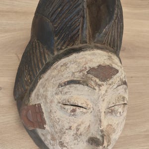 May include: A carved wooden mask featuring a white-pigmented face with closed eyes, a narrow nose, and a small mouth. The top of the mask has a dark brown, textured, fan-like crest, while the sides display reddish-brown ear-like ornaments.