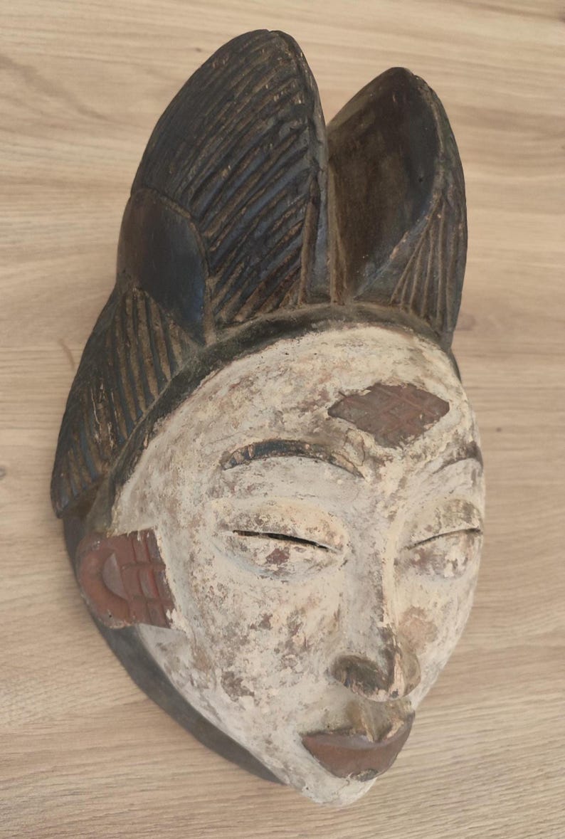 May include: A carved wooden mask featuring a white-pigmented face with closed eyes, a narrow nose, and a small mouth. The top of the mask has a dark brown, textured, fan-like crest, while the sides display reddish-brown ear-like ornaments.