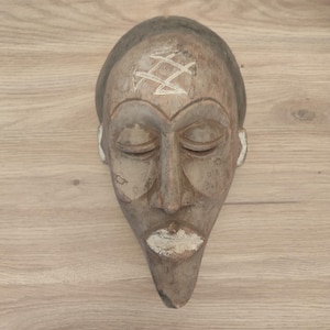 May include: A carved wooden mask with a pointed chin and closed eyes. The mask features a weathered brown finish with white pigment accents on the forehead, ears, and lips. A distinct white geometric symbol is etched into the center of the forehead.
