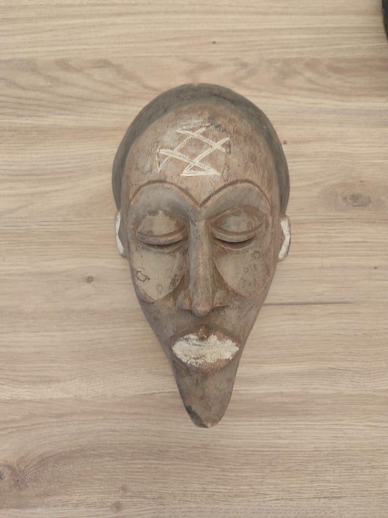 May include: A carved wooden mask with a pointed chin and closed eyes. The mask features a weathered brown finish with white pigment accents on the forehead, ears, and lips. A distinct white geometric symbol is etched into the center of the forehead.