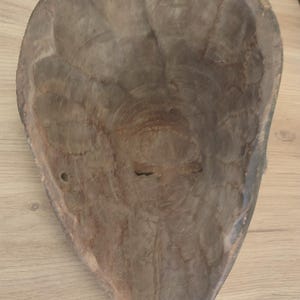 May include: A hollowed-out, teardrop-shaped wooden mask viewed from the interior. The surface shows visible chisel marks and a natural, light brown wood grain texture. The structure is concave, tapering to a point at the bottom edge.