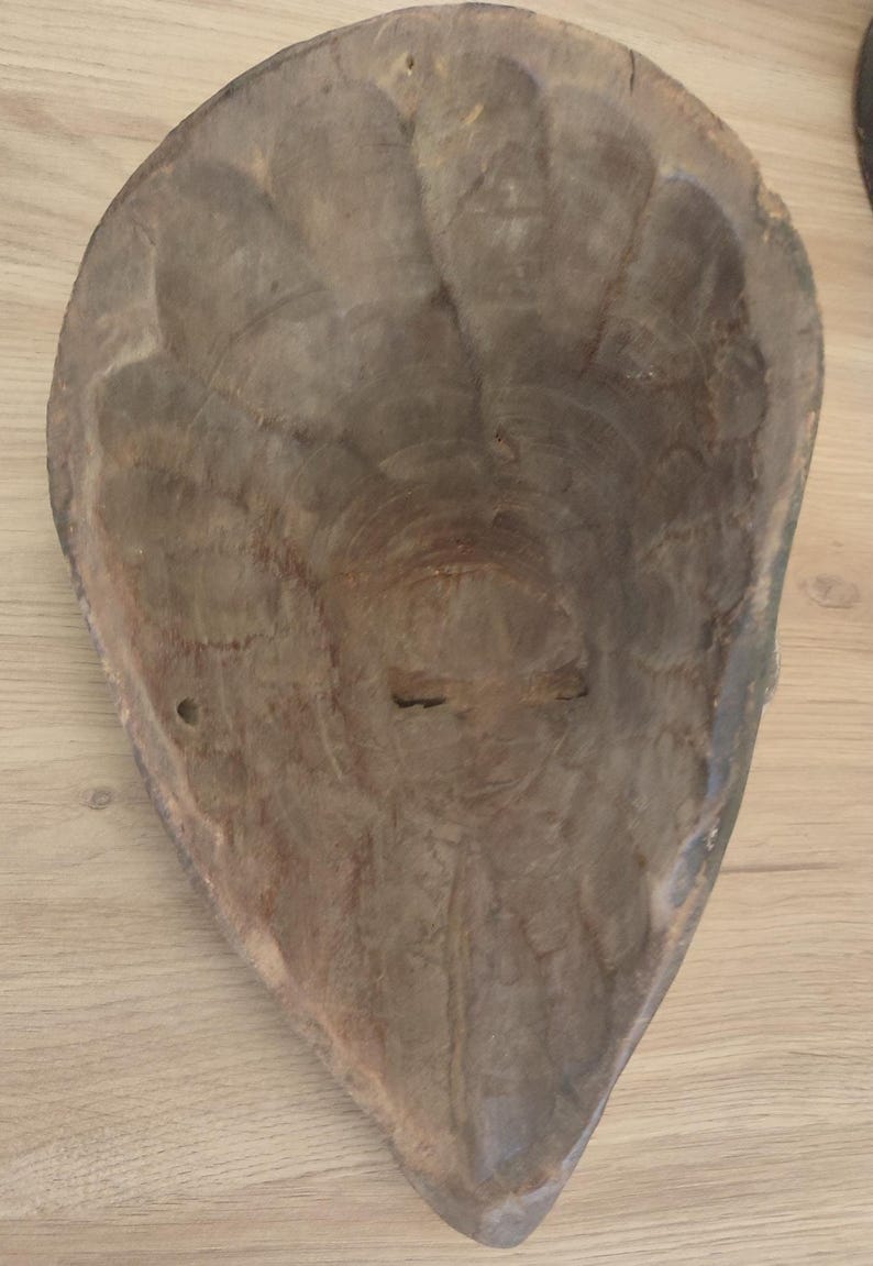 May include: A hollowed-out, teardrop-shaped wooden mask viewed from the interior. The surface shows visible chisel marks and a natural, light brown wood grain texture. The structure is concave, tapering to a point at the bottom edge.