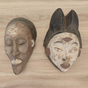 May include: Two carved wooden masks. The left mask is brown with a white geometric symbol on the forehead and white accents on the lips. The right mask features a dark brown textured headdress, a white face, and dark brown lips and markings.