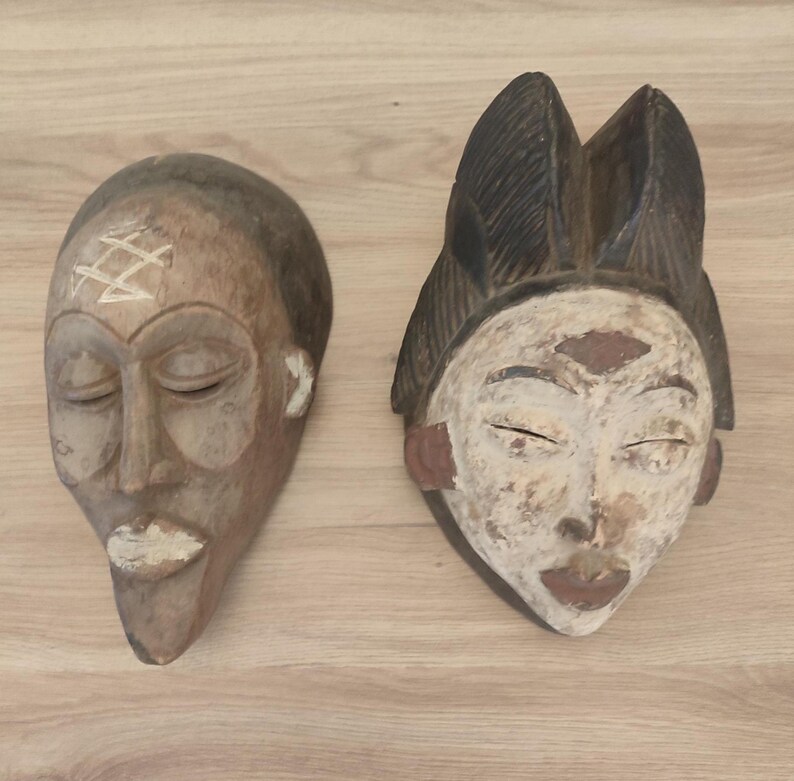 May include: Two carved wooden masks. The left mask is brown with a white geometric symbol on the forehead and white accents on the lips. The right mask features a dark brown textured headdress, a white face, and dark brown lips and markings.