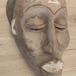 May include: A carved wooden mask with a weathered brown finish, featuring closed eyes, a prominent nose, and a pointed chin. The forehead displays a white geometric symbol, while the lips and ear details are accented with white pigment.