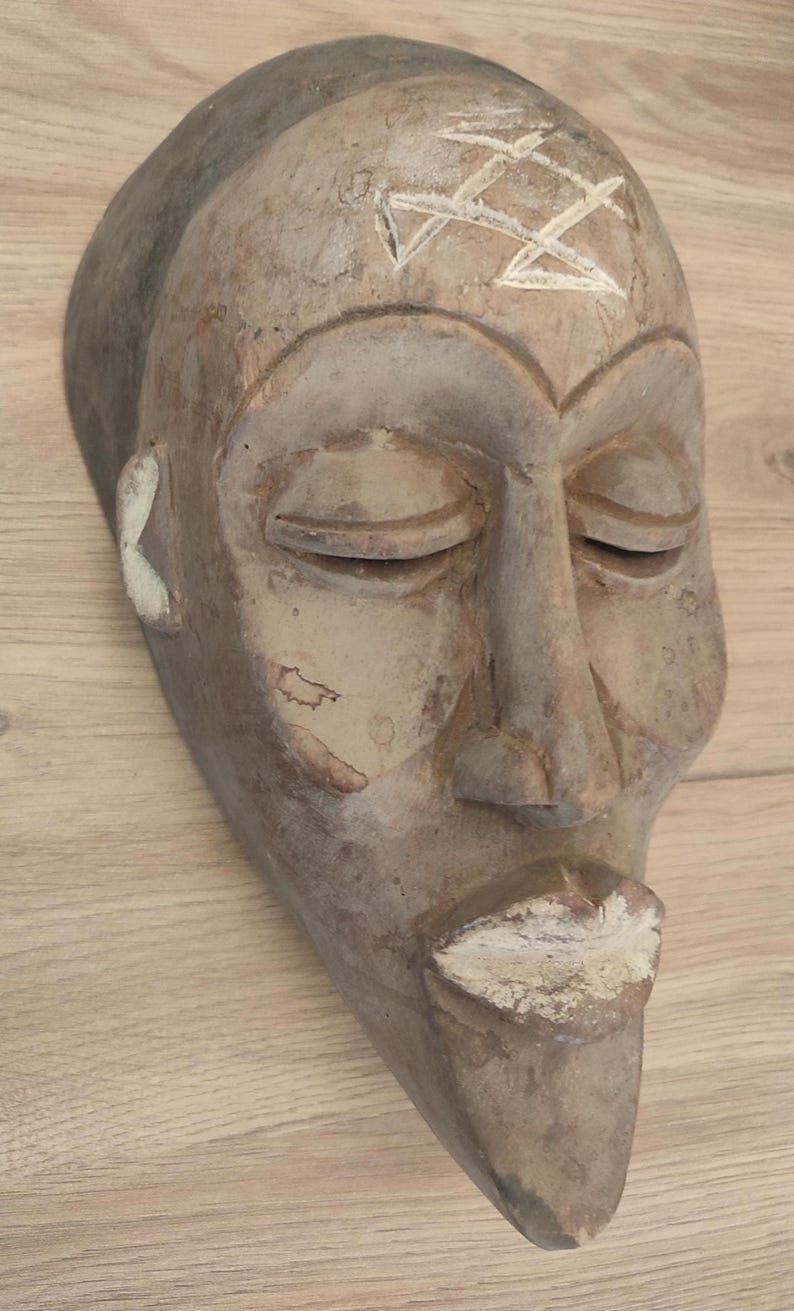 May include: A carved wooden mask with a weathered brown finish, featuring closed eyes, a prominent nose, and a pointed chin. The forehead displays a white geometric symbol, while the lips and ear details are accented with white pigment.