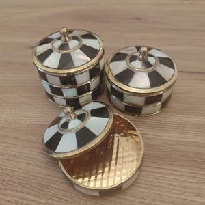 May include: Three round containers with lids, featuring a black and white checkered pattern made of inlaid material. Each container has a gold-toned metal rim and a small gold-toned knob on the center of the lid. One lid is removed and placed beside its base.