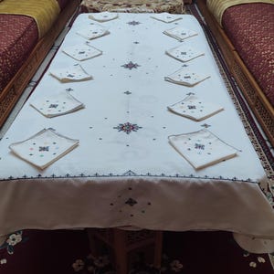 May include: White tablecloth with embroidered floral designs and a set of white napkins arranged on top. The tablecloth is laid out on a table with a red and white patterned rug underneath.