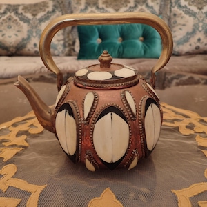 Könnte beinhalten: Eine Kupfer-Teekanne mit Messinggriff und Ausguss. Der Korpus weist weiße und schwarze ovale Designs auf. Der Deckel ist weiß mit schwarzen Punkten. Die Teekanne steht auf einer dekorativen, goldfarbenen Tischdecke.