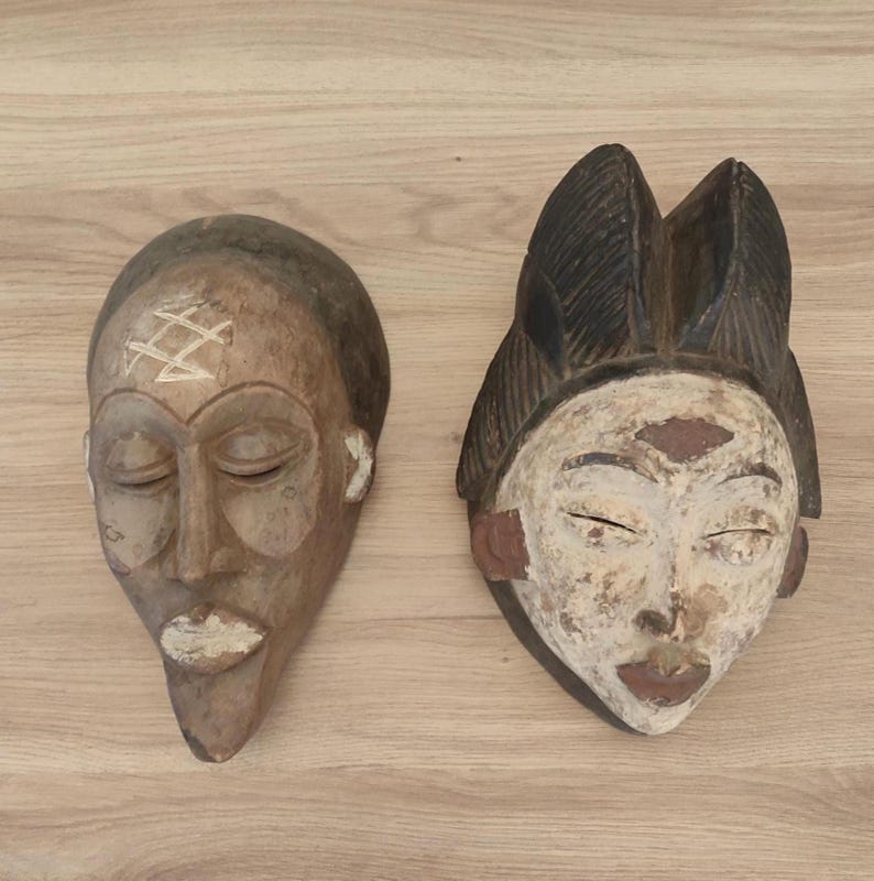 May include: Two carved wooden masks. The left mask is brown with a light-colored geometric symbol on the forehead and white paint on the lips. The right mask features a dark brown textured headdress, a white-painted face, and dark brown lips.