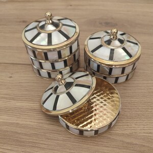 May include: Three round containers with lids, featuring a radial pattern of white mother-of-pearl and black segments framed by gold-toned metal edges. Each lid has a small gold-toned knob at the centre, and the interior of the open container is gold.