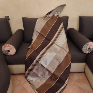 May include: A brown and tan striped scarf draped over a dark grey sofa. The scarf has a patchwork design with varying shades of brown and tan. The sofa has two cylindrical throw pillows with floral patterns.