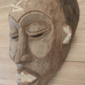 May include: A hand-carved wooden mask featuring a smooth, light brown finish with a dark brown forehead. The mask displays closed eyes, a prominent nose, and full lips highlighted with white pigment, plus a white geometric symbol on the forehead.