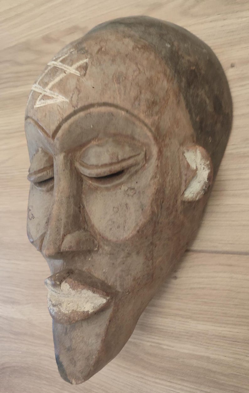 May include: A hand-carved wooden mask featuring a smooth, light brown finish with a dark brown forehead. The mask displays closed eyes, a prominent nose, and full lips highlighted with white pigment, plus a white geometric symbol on the forehead.
