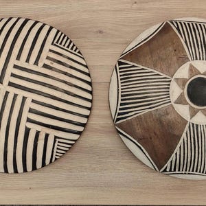 May include: Two circular wooden wall plaques featuring hand-carved geometric patterns. The left plaque displays a black and white striped grid design. The right plaque features a central black circle surrounded by a star and radiating brown and white lines.