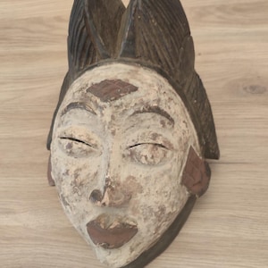 May include: A carved wooden mask featuring a weathered white finish on the face, dark brown lips, and a prominent dark brown headdress with vertical ridges. The mask displays a diamond-shaped carving on the forehead and textured side panels.