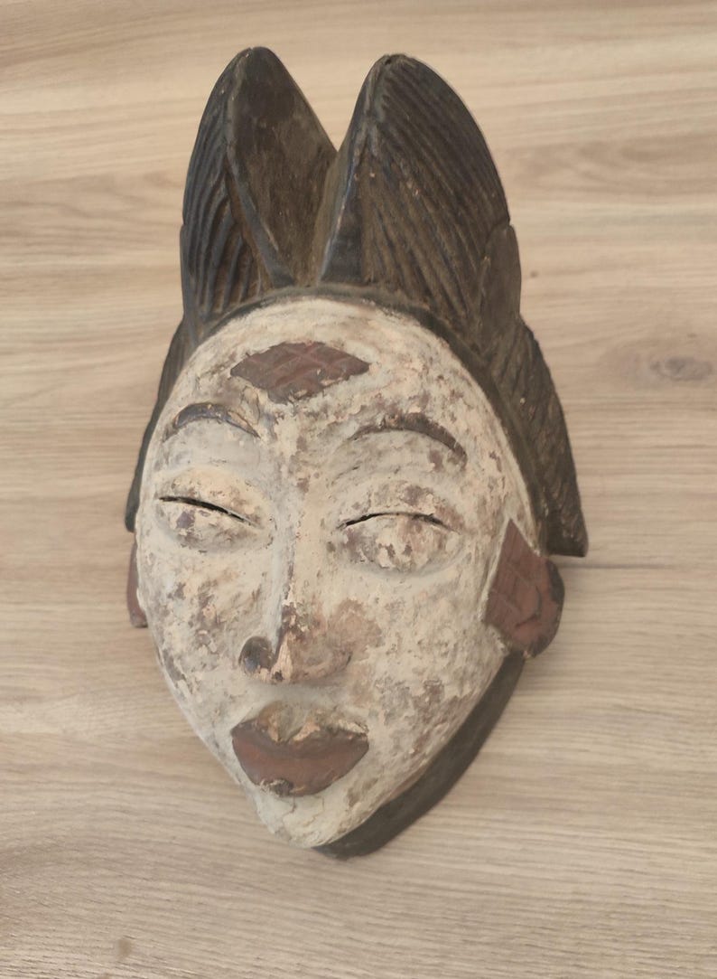 May include: A carved wooden mask featuring a weathered white finish on the face, dark brown lips, and a prominent dark brown headdress with vertical ridges. The mask displays a diamond-shaped carving on the forehead and textured side panels.