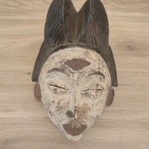 May include: A carved wooden mask featuring a weathered off-white face with dark brown accents on the lips, ears, and forehead. The mask has a prominent, textured dark brown headdress with vertical ridges and closed, almond-shaped eyes.