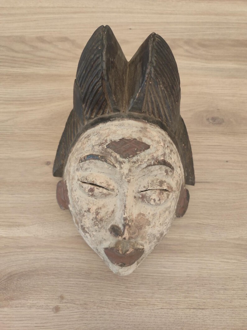 May include: A carved wooden mask featuring a weathered off-white face with dark brown accents on the lips, ears, and forehead. The mask has a prominent, textured dark brown headdress with vertical ridges and closed, almond-shaped eyes.
